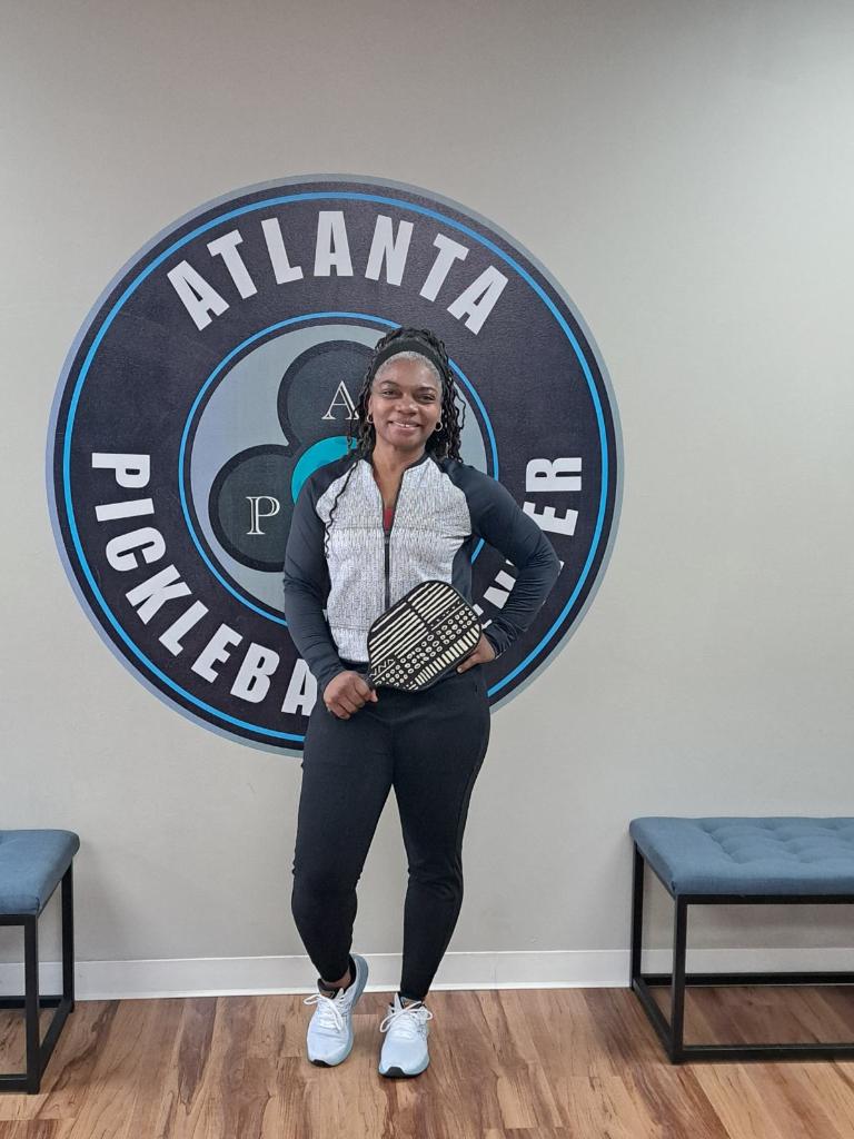 A woman posing in front of a large 'Atlanta Pickleball Club' logo on a wall, holding an African print pickleball paddle that she purchased from Pickleblack.
